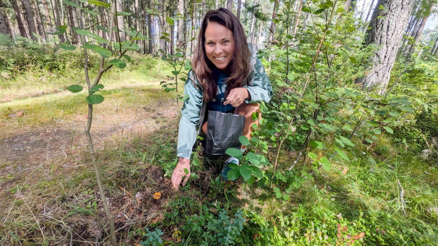 En kvinna sitter på huk i ett skogsområde och ler mot kameran samtidigt som hon pekar på en liten orange svamp som ligger på marken bland gröna växter.