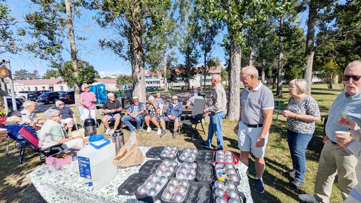 En grupp människor sitter och står utomhus i en park nära ett bord med drycker och brickor med ägg en solig dag. I bakgrunden syns träd och byggnader.