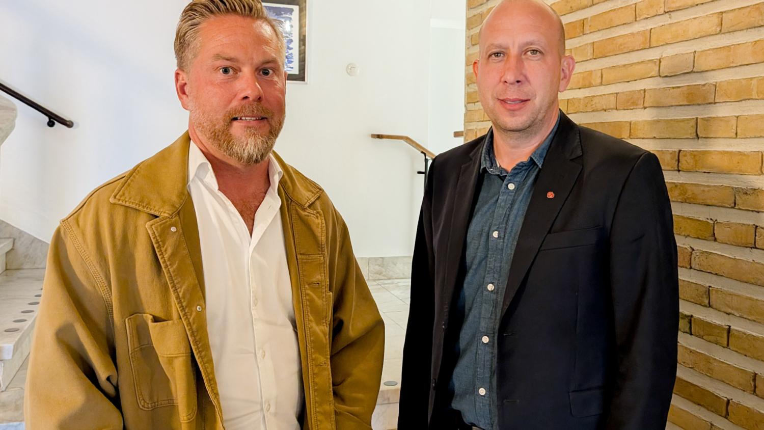 Två män står inomhus nära en trappa och en tegelvägg, båda vända mot kameran; den ene bär en beige jacka, den andre en mörk kavaj.