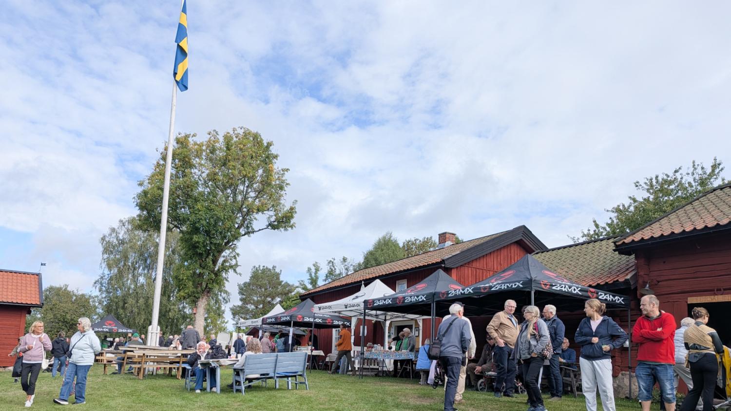 Människor samlas vid ett utomhusarrangemang med matstånd, träbyggnader och en hissad svensk flagga på en gräsplan under en molnig himmel.