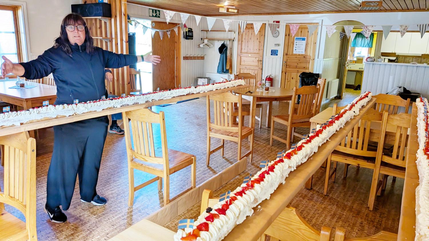 En person står med utsträckta armar bredvid en lång efterrätt toppad med vispad grädde och jordgubbar, som står uppställd på träbord i ett festligt dekorerat rum.