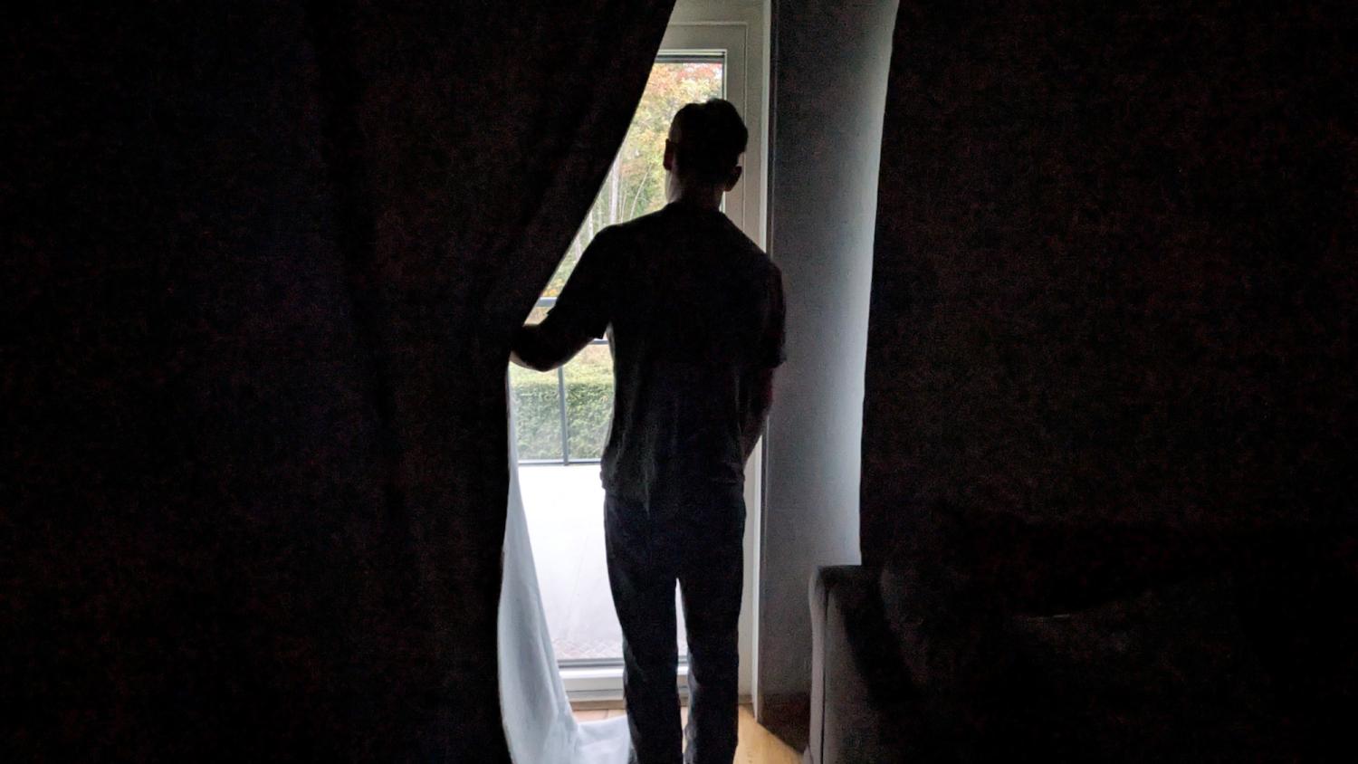 En person står inomhus och håller delvis undan en gardin medan hen tittar ut genom en glasdörr mot en balkong.