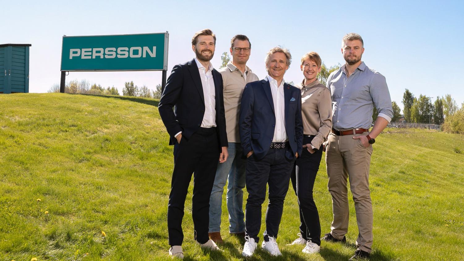 Fem personer står utomhus på gräset nära en stor skylt med texten ”PERSSON”, med träd och en klar himmel i bakgrunden.