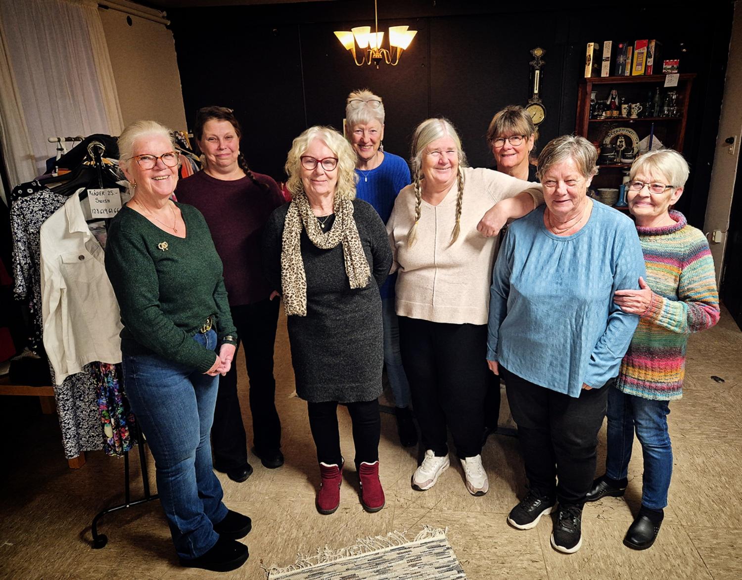 Åtta äldre kvinnor står tillsammans inomhus och ler mot kameran. Klädhängare och hyllor syns i bakgrunden. Miljön verkar avslappnad och vänlig.