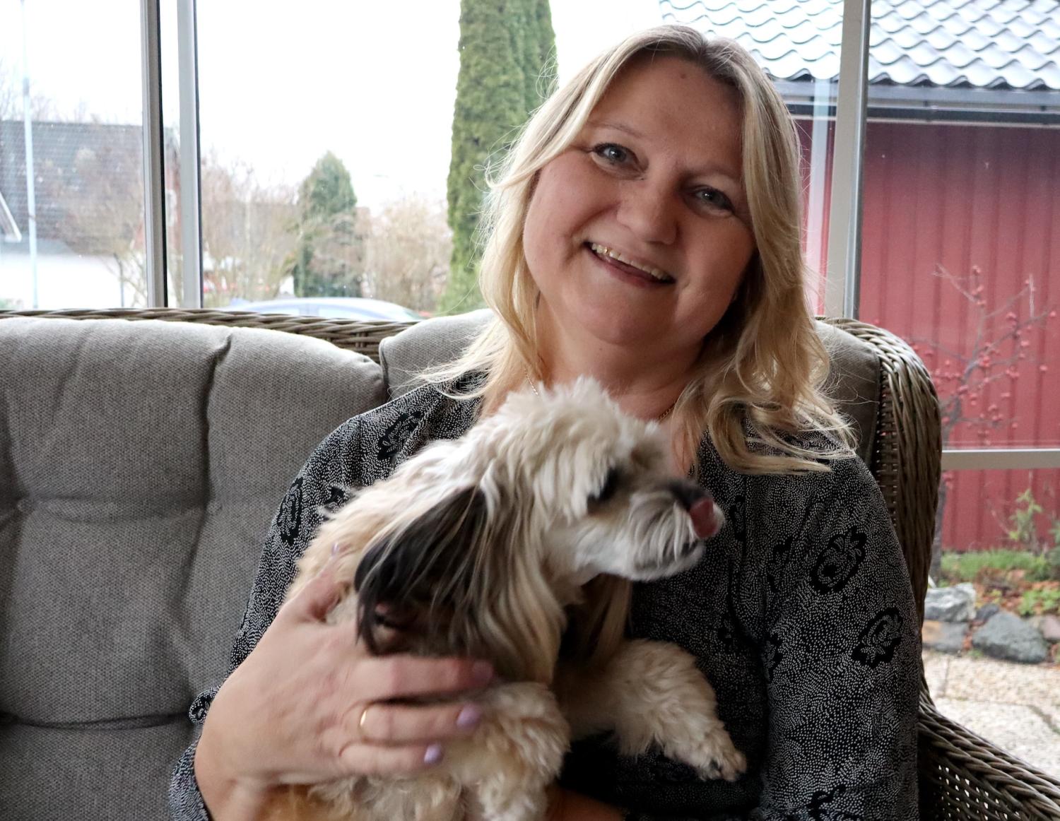 En kvinna som sitter på en soffa håller i en liten hund. Hon ler och det finns ett stort fönster bakom henne som visar en utomhusvy.