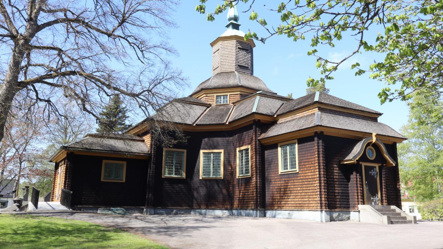 En träkyrka med ett sexkantigt torn och flera spetsiga tak, omgiven av träd och en gräsyta en solig dag.