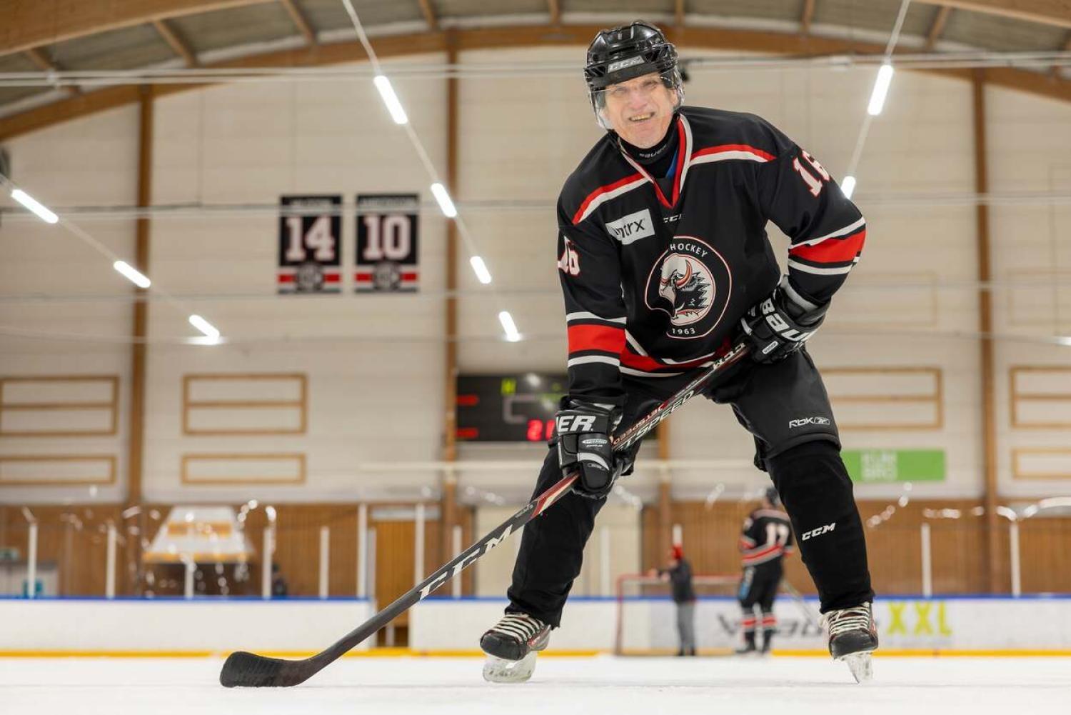 En hockeyspelare i svart och röd uniform poserar på isbanan, håller i en hockeyklubba och ler mot kameran. Andra spelare och banderoller syns i bakgrunden.