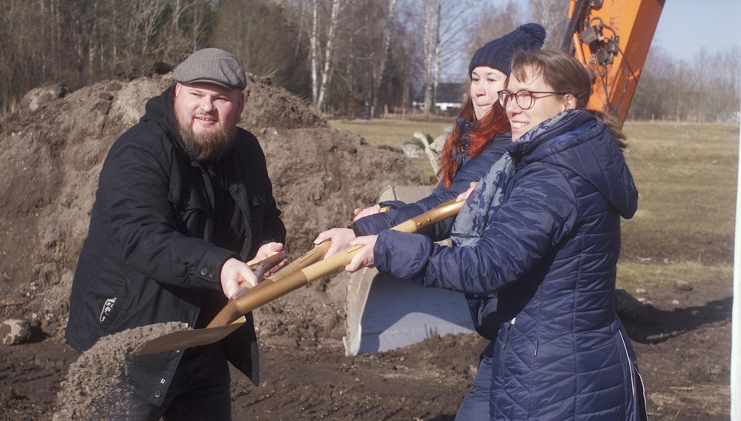 Tre individer som deltar i ett ceremoniellt banbrytande med spadar, med en grävmaskin i bakgrunden.
