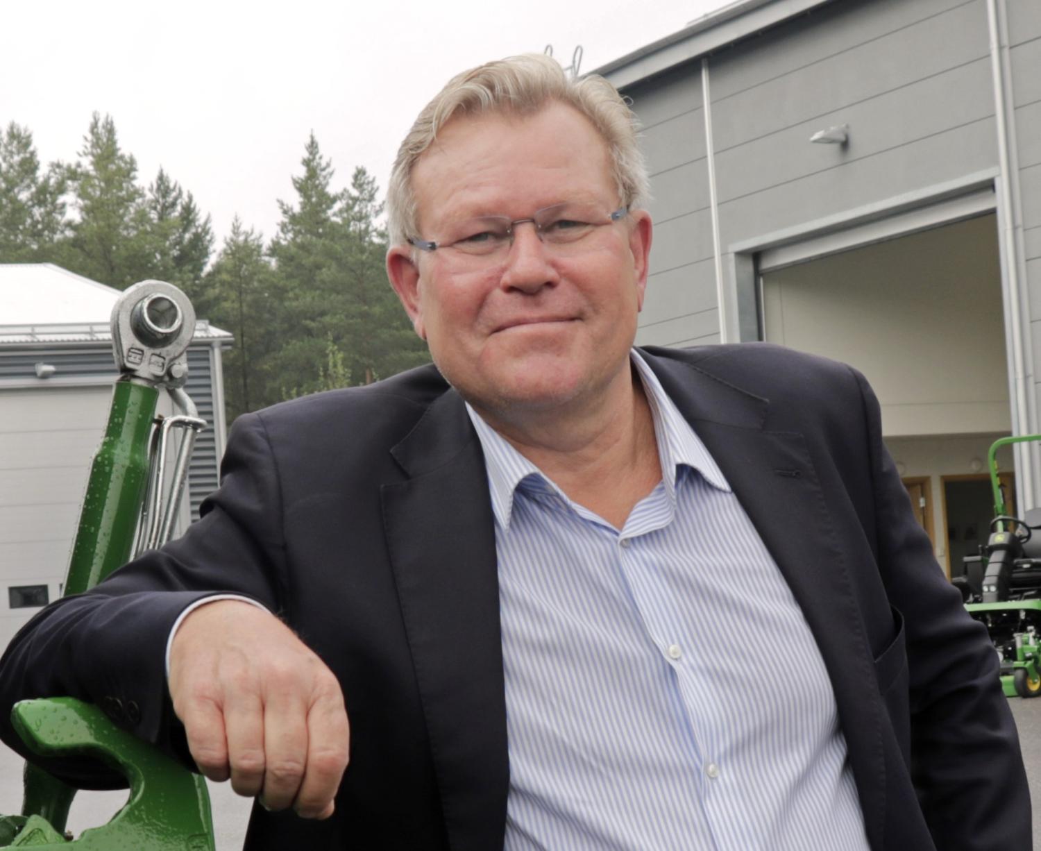 En man i kostym och glasögon står utomhus och lutar sig mot en grön maskin framför en modern industribyggnad, med träd i bakgrunden.