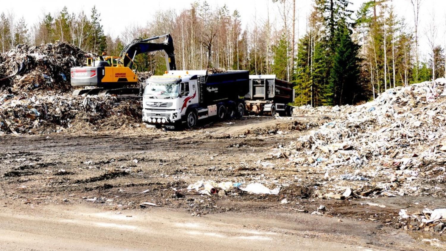En stor lastbil och en grävmaskin arbetar bland avfallshögar på en utomhusdeponi, med träd synliga i bakgrunden.