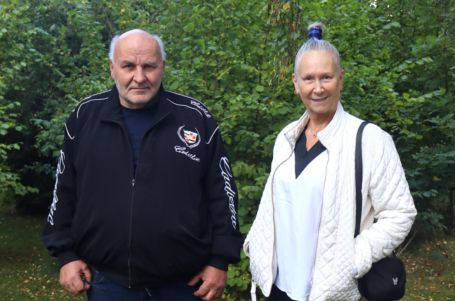 En äldre man och kvinna står utomhus framför grönt lövverk. Mannen bär en svart jacka, och kvinnan bär en vit vadderad jacka och har håret uppsatt i en knut.
