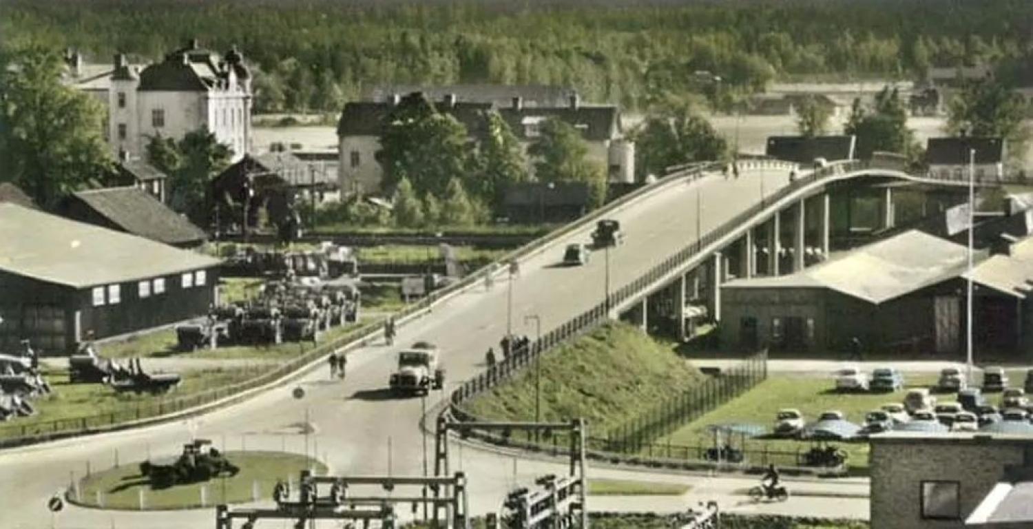 Ett färglagt historiskt foto visar en bro över en väg med bilar, omgiven av byggnader, parkerade fordon, träd och en flod i bakgrunden.