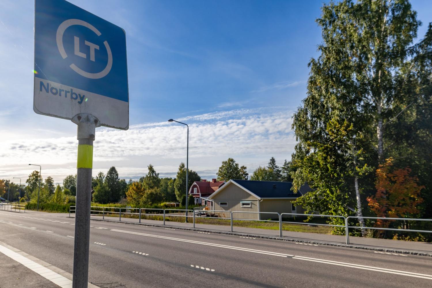 En busshållplatsskylt vid vägen med texten ”Norrby”, med träd, hus och en klarblå himmel i bakgrunden.
