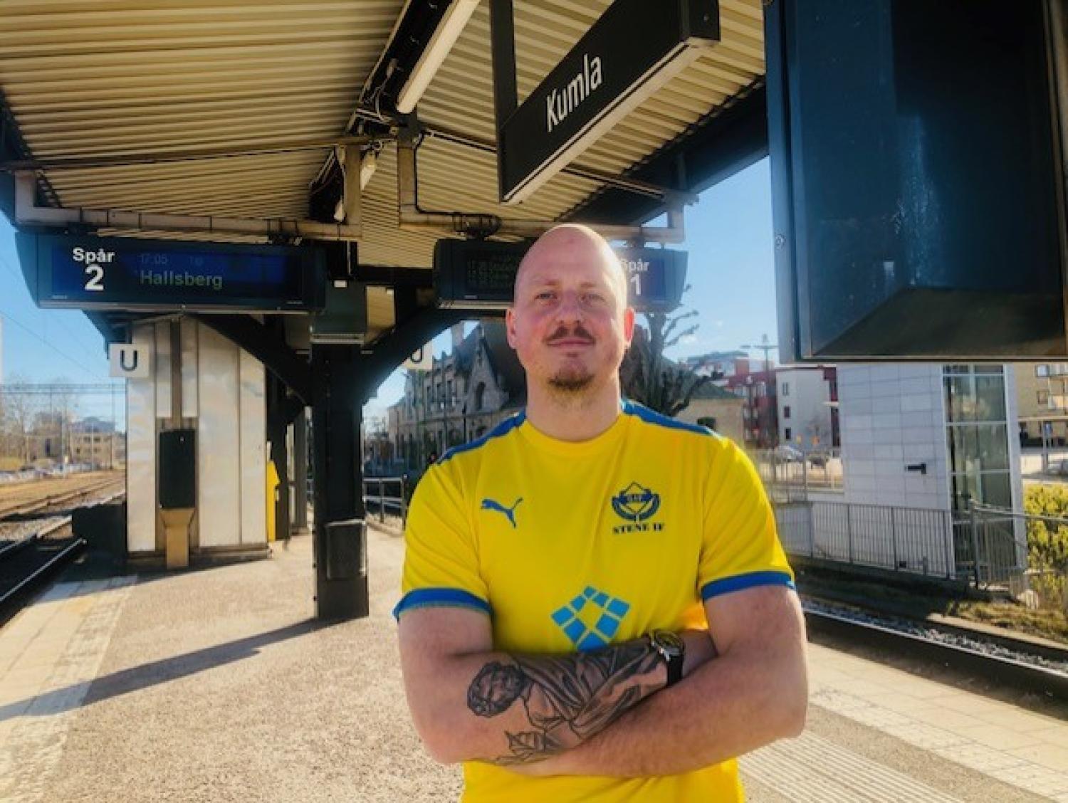 En person står på en järnvägsstation perrong iförd en gul sporttröja. Skyltar för spår och stationsnamnet