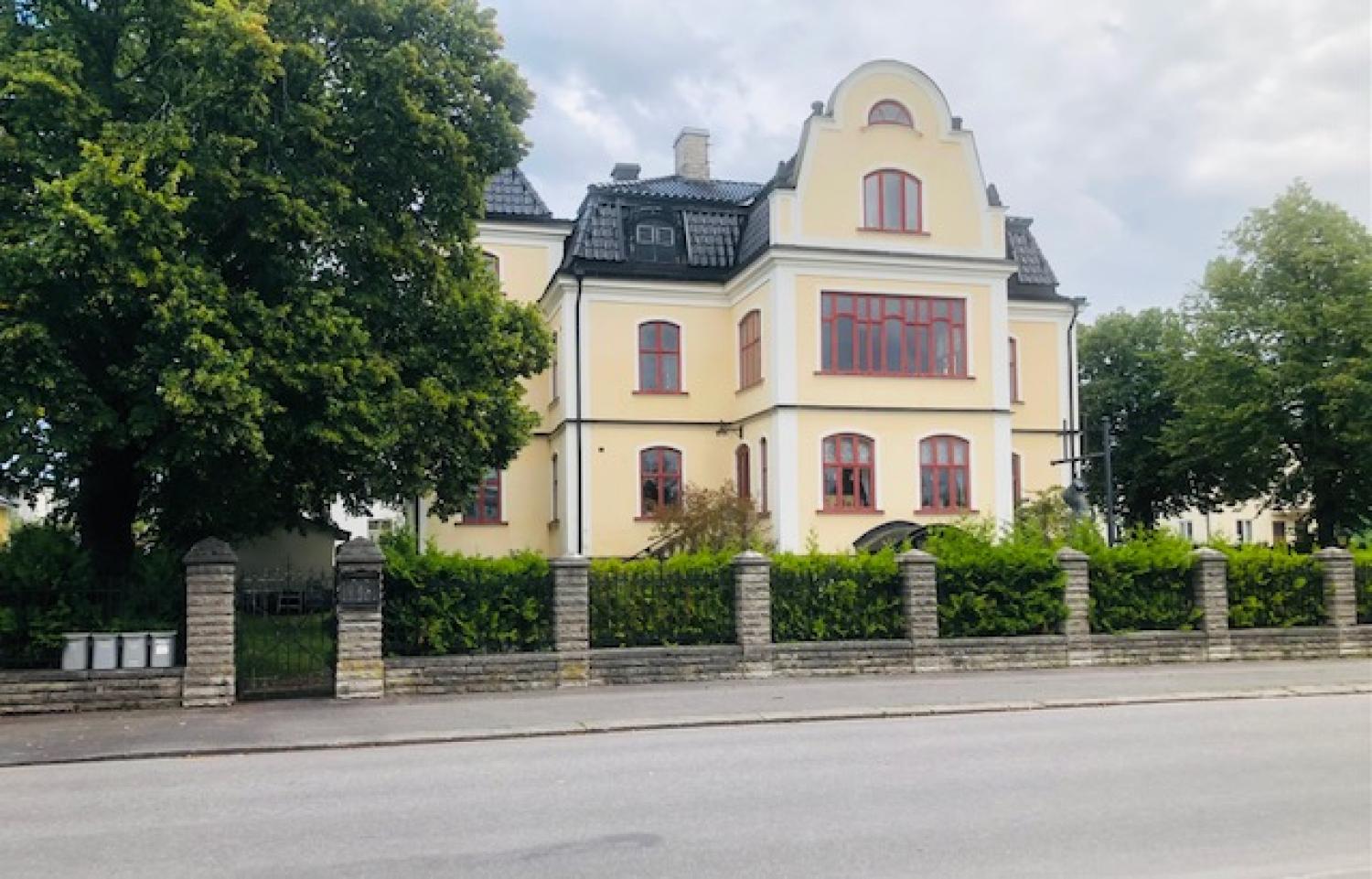 En stor gul byggnad med rödmålade fönster står bakom ett stengärde och gröna träd en molnig dag.