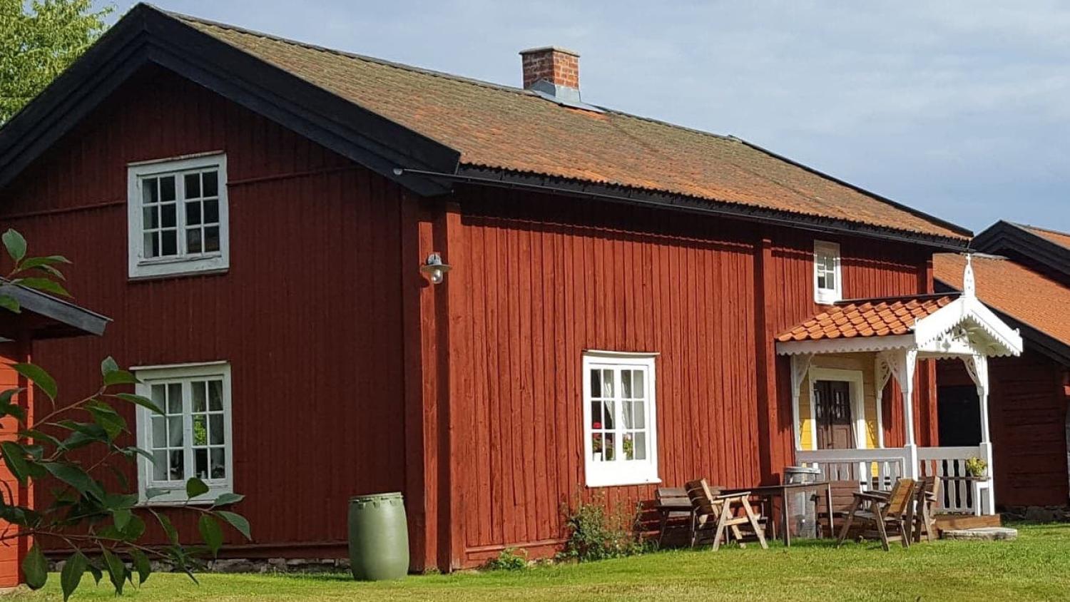 Ett traditionellt rött trähus med vita detaljer, en liten veranda, utemöbler och en grön trädgård en solig dag.