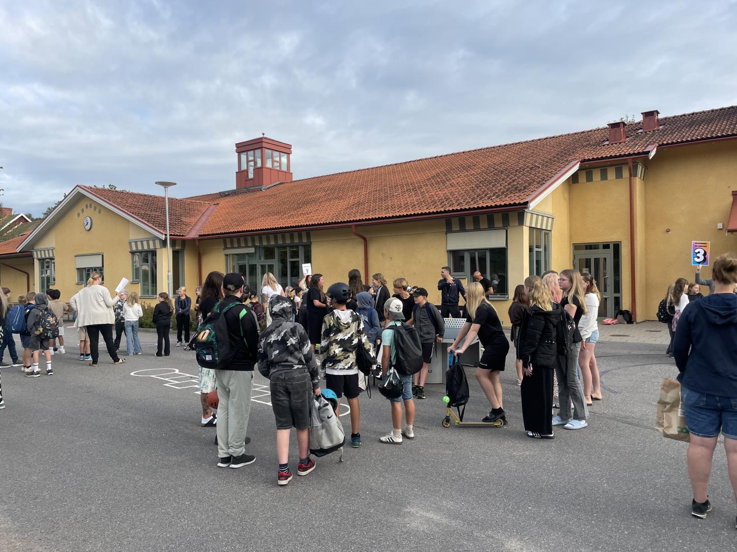 En grupp människor, däribland barn och vuxna, samlas utanför en gul byggnad med rött tak en molnig dag.