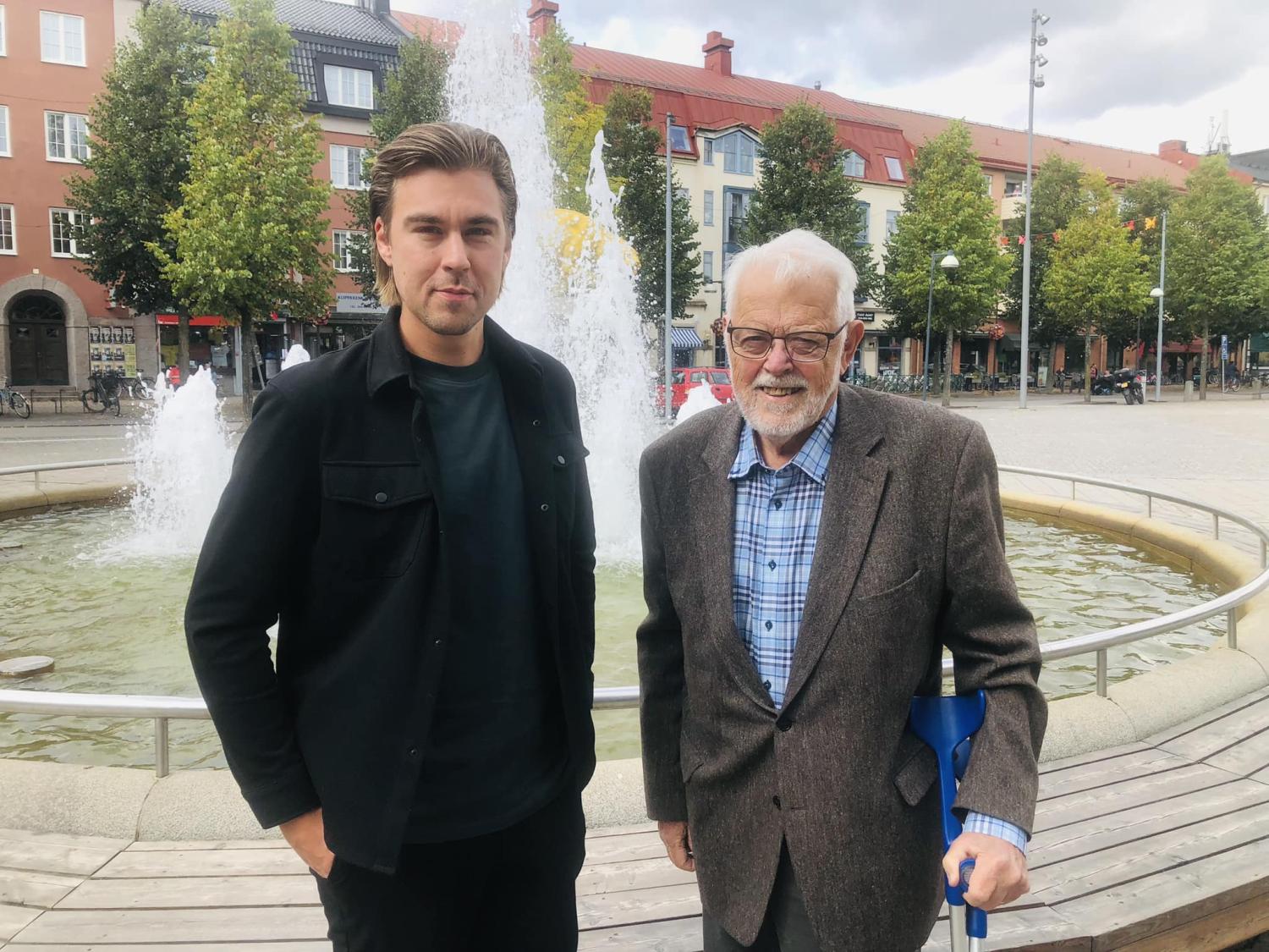 Två män står framför en fontän på ett stadstorg med byggnader och träd i bakgrunden.