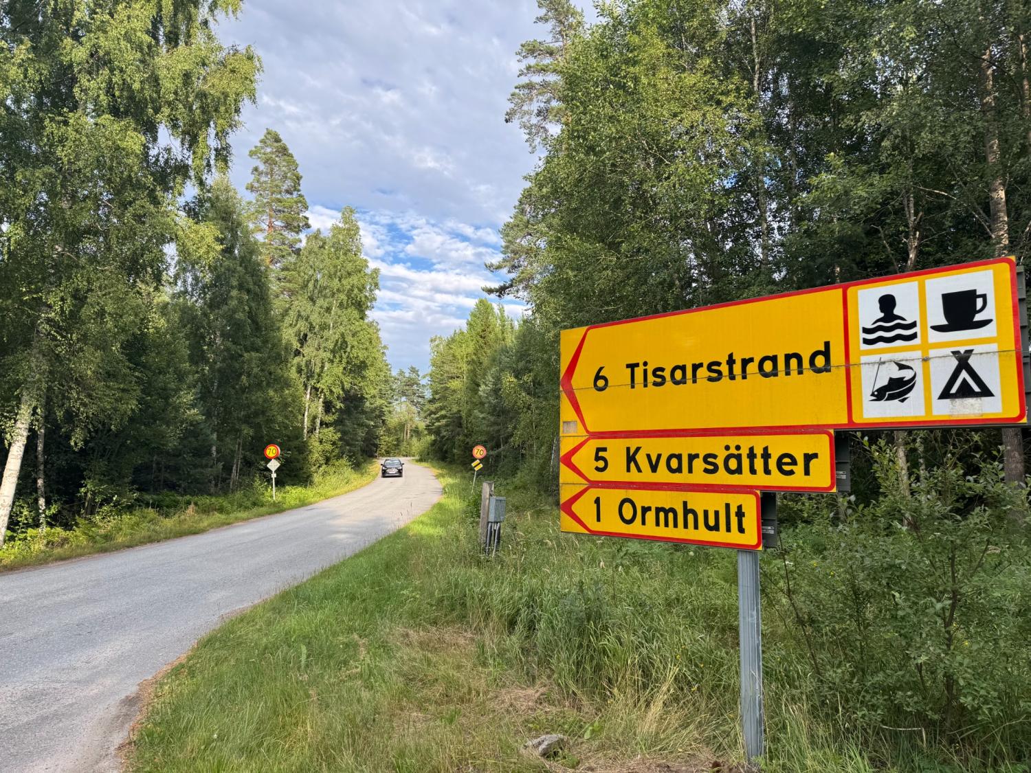 Gula vägskyltar vid en skogsväg pekar mot Tisarsstrand, Kvarsätter och Ormhult; symboler anger att det finns bad, camping och matställen i närheten. En bil kör iväg i fjärran.