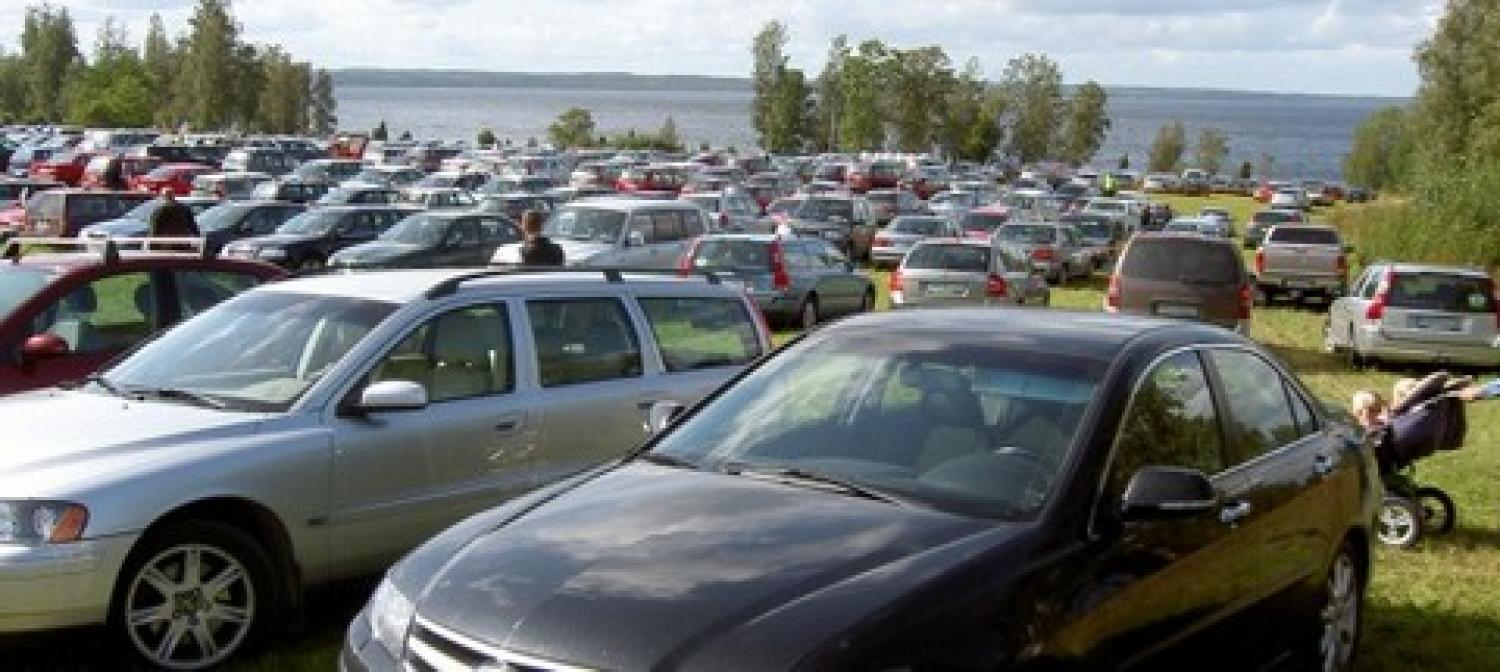 Ett stort gräsbevuxet fält som används som parkeringsplats, fyllt med parkerade bilar, med en sjö och träd i bakgrunden.