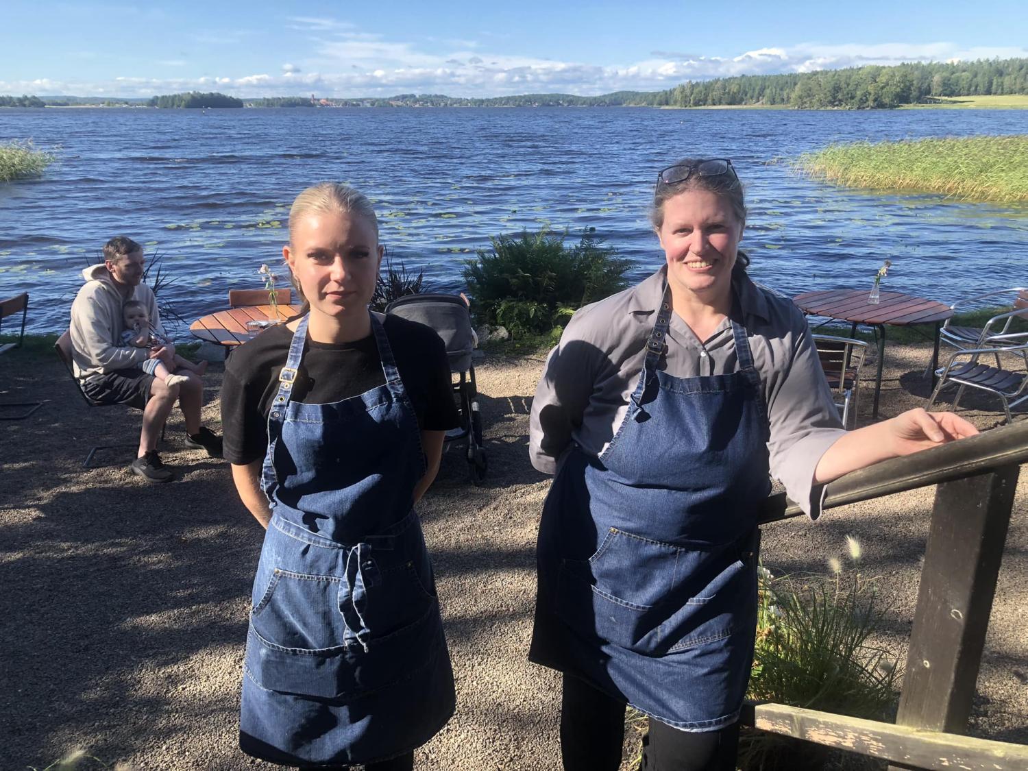 Två personer som bär blå förkläden står utomhus vid en sjö. Bord och stolar är placerade på grusmarken och en annan person sitter i bakgrunden nära sjön. Träd och klar himmel syns.
