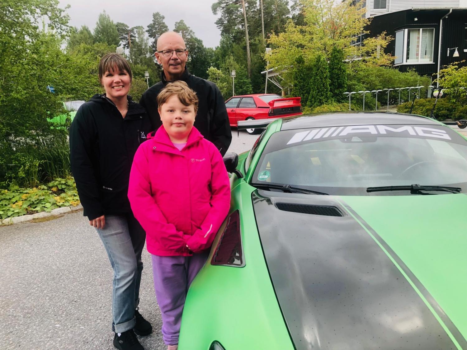 Tre personer står bredvid en grön AMG-bil som står parkerad på en bostadsgata, med grönska och andra fordon i bakgrunden.