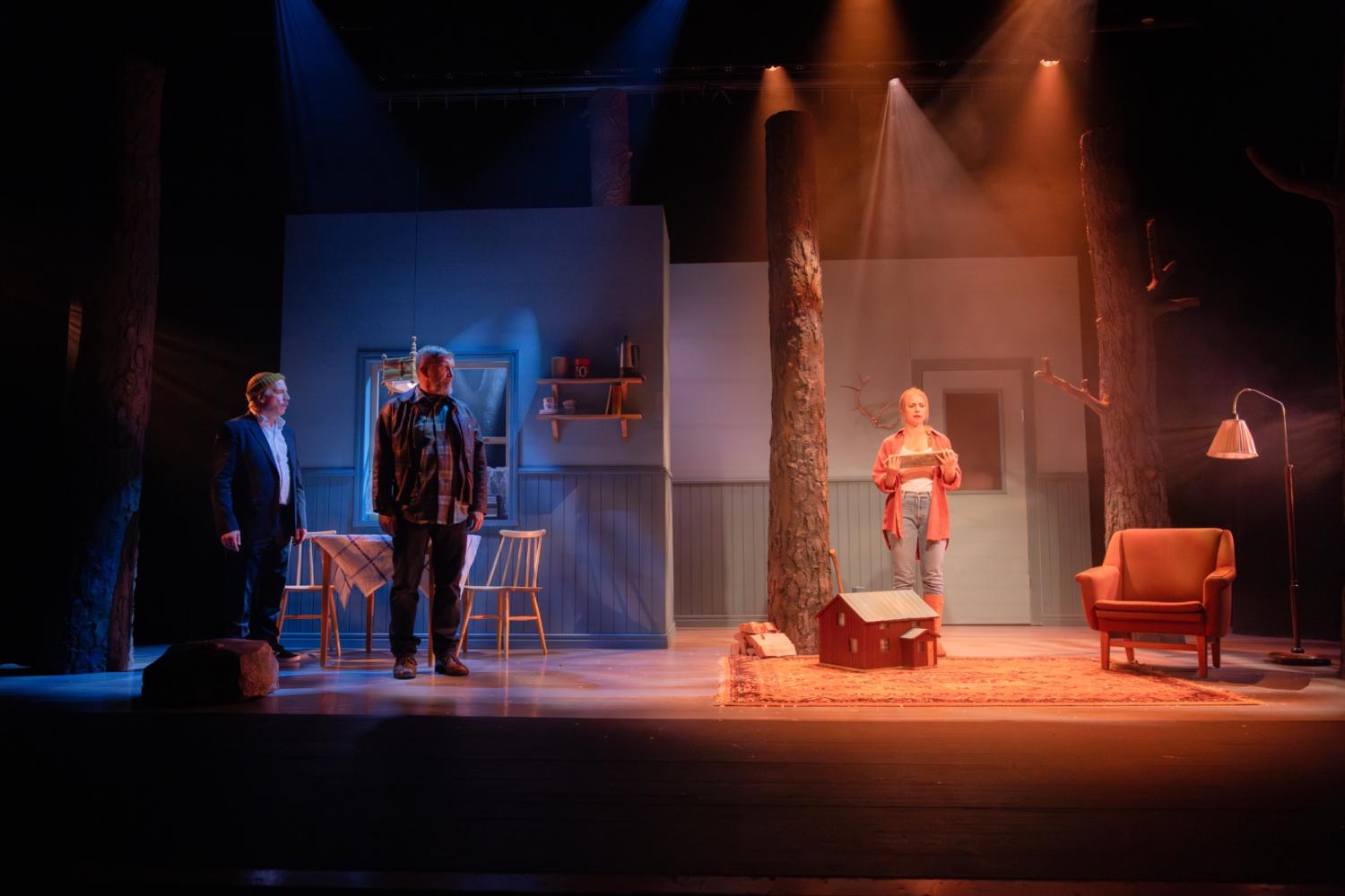 Tre personer står på en scen som liknar ett vardagsrum med träd, möbler och dramatisk belysning, vilket antyder en scen från en teaterpjäs.