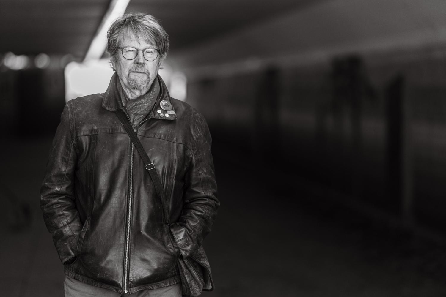 En äldre man med glasögon och skägg står i en gångtunnel, iklädd en skinnjacka och bär en axelremsväska. Bilden är i svartvitt.