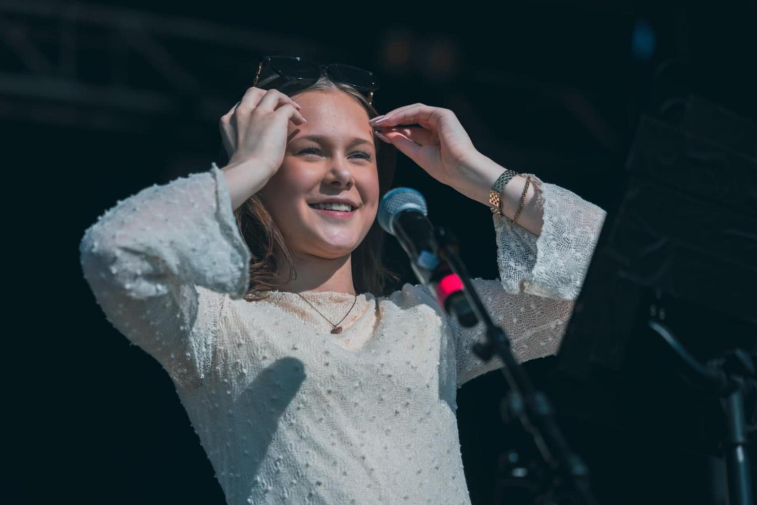 En ung kvinna i en vit strukturerad klänning står vid en mikrofon på scenen, ler och justerar sina solglasögon.
