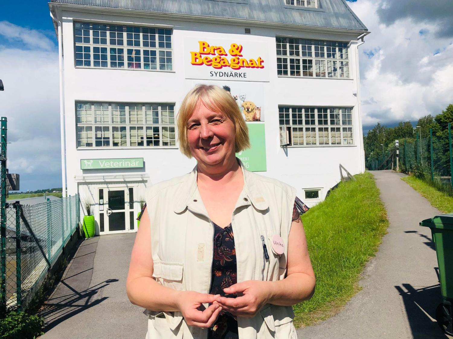 En person står och ler framför en vit byggnad med skylten