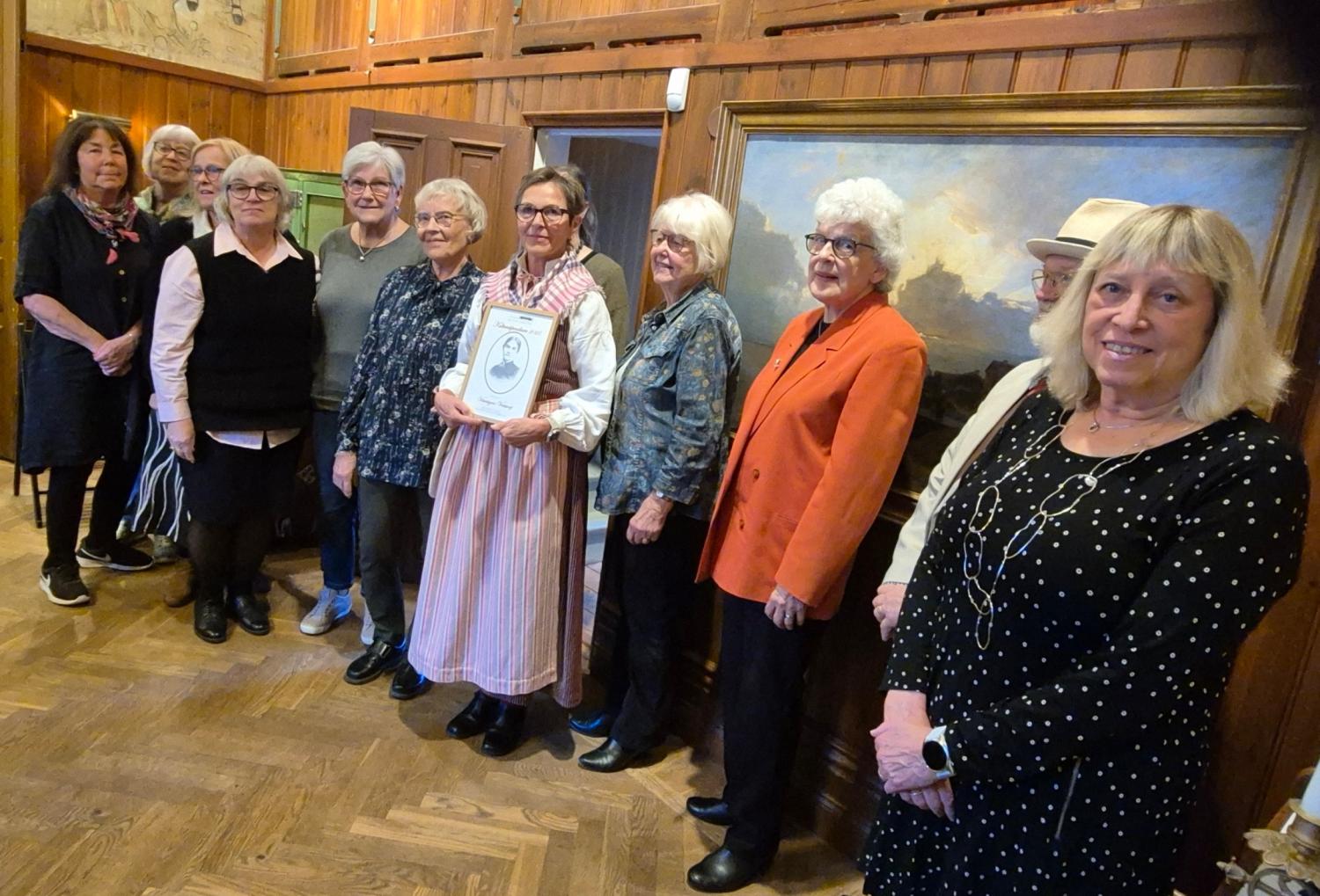 En grupp på elva äldre vuxna står inomhus i en rad, med en person i mitten som håller ett inramat certifikat. Målningar pryder de träpanelklädda väggarna bakom dem.