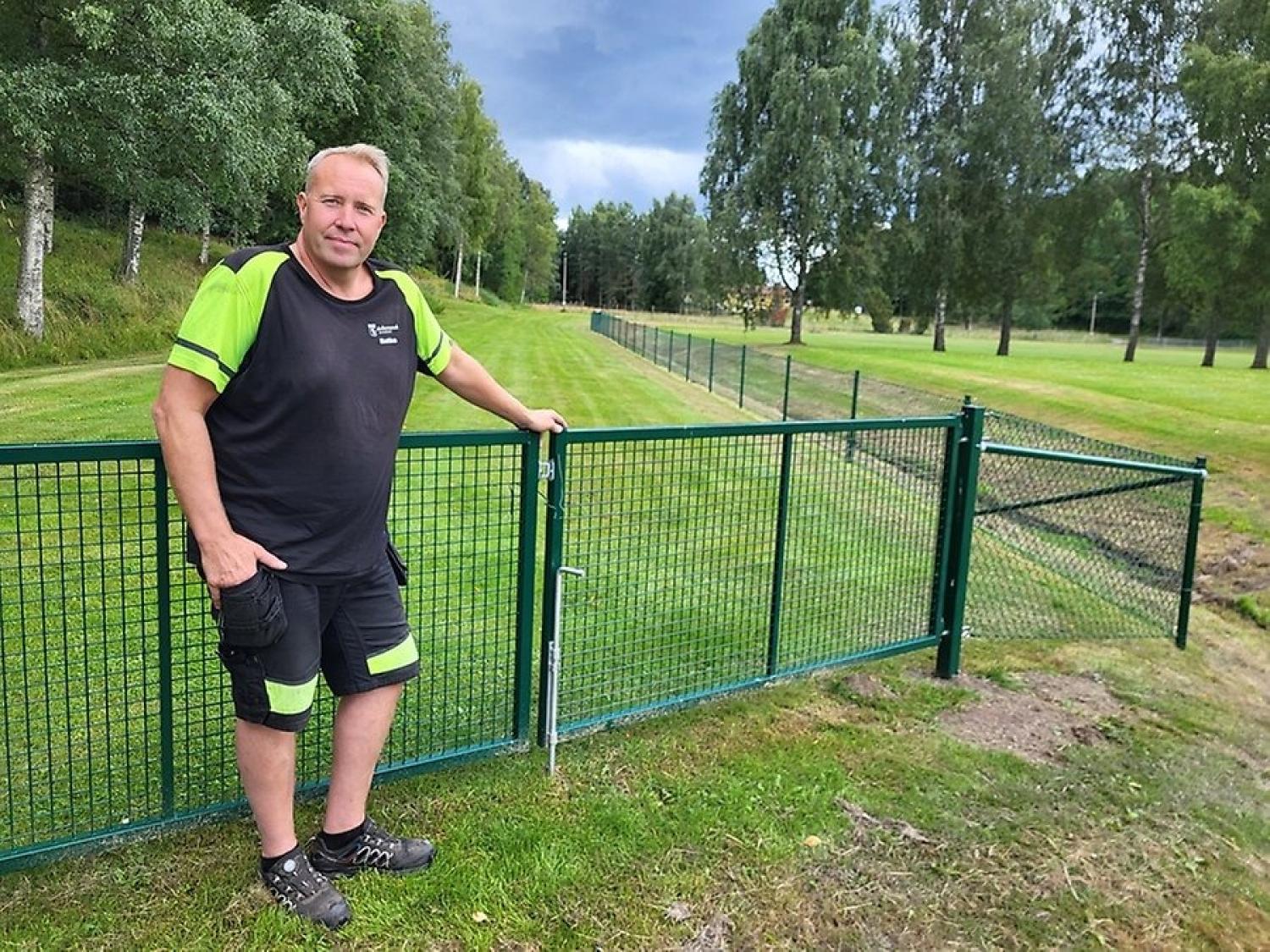 En man i arbetskläder står bredvid ett grönt metallstaket på en gräsbevuxen äng med träd i bakgrunden under en molnig himmel.