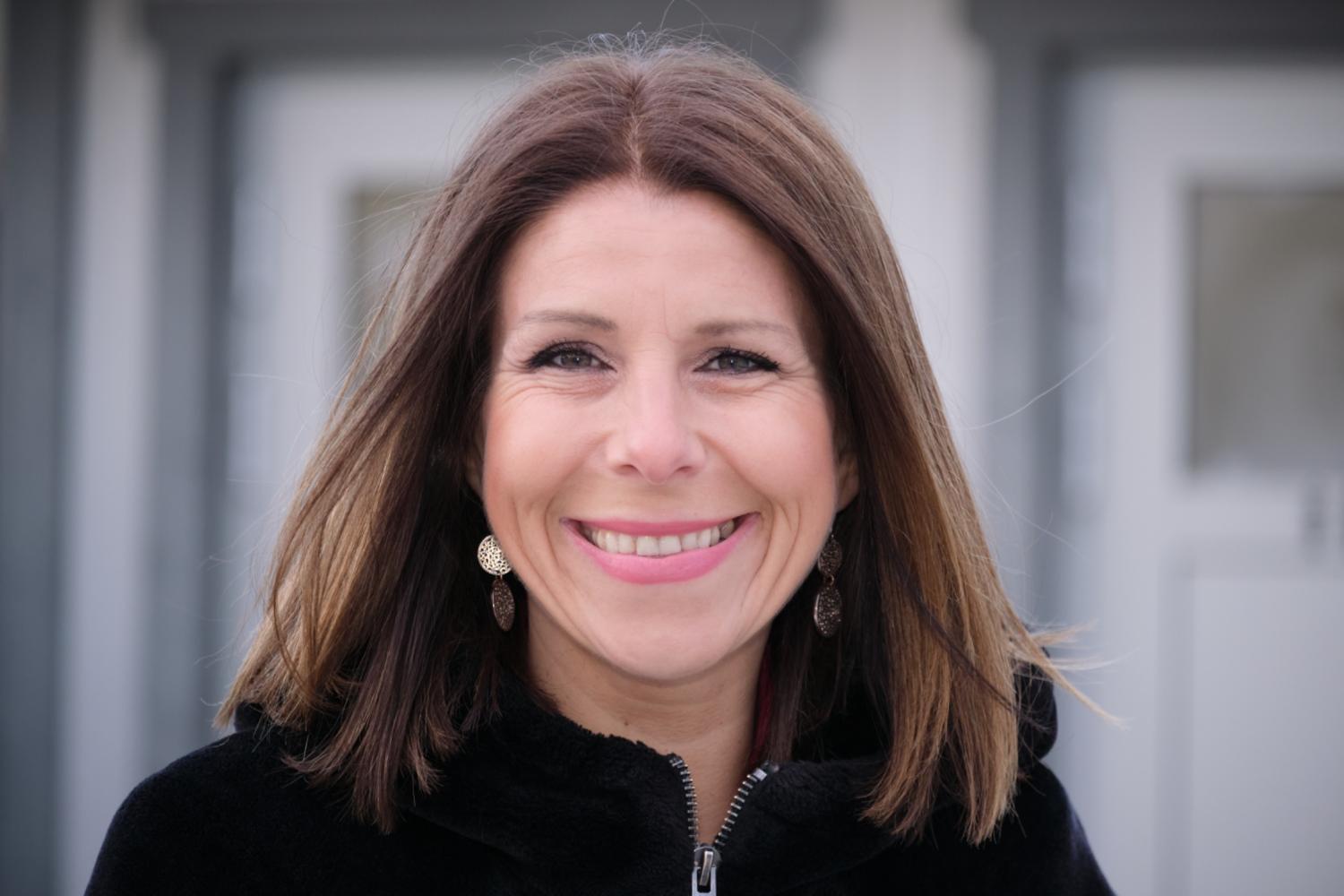 En kvinna med rakt brunt hår, klädd i svart kappa och bladformade örhängen, ler utanför en byggnad.