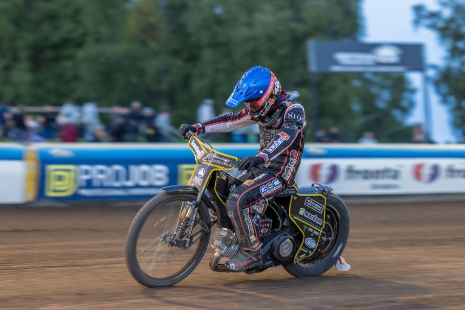 En motorcykelförare iförd full utrustning och blå hjälm kör en motorcykel på en grusbana under en speedwaytävling, med åskådare i bakgrunden.