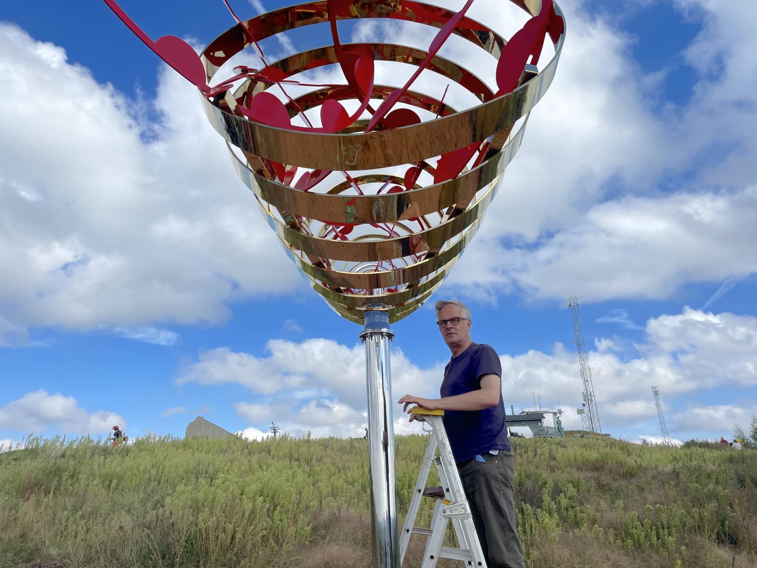 En man står på en stege bredvid en stor utomhusskulptur i metall med röda element, placerad på en gräsmatta under en delvis molnig himmel.