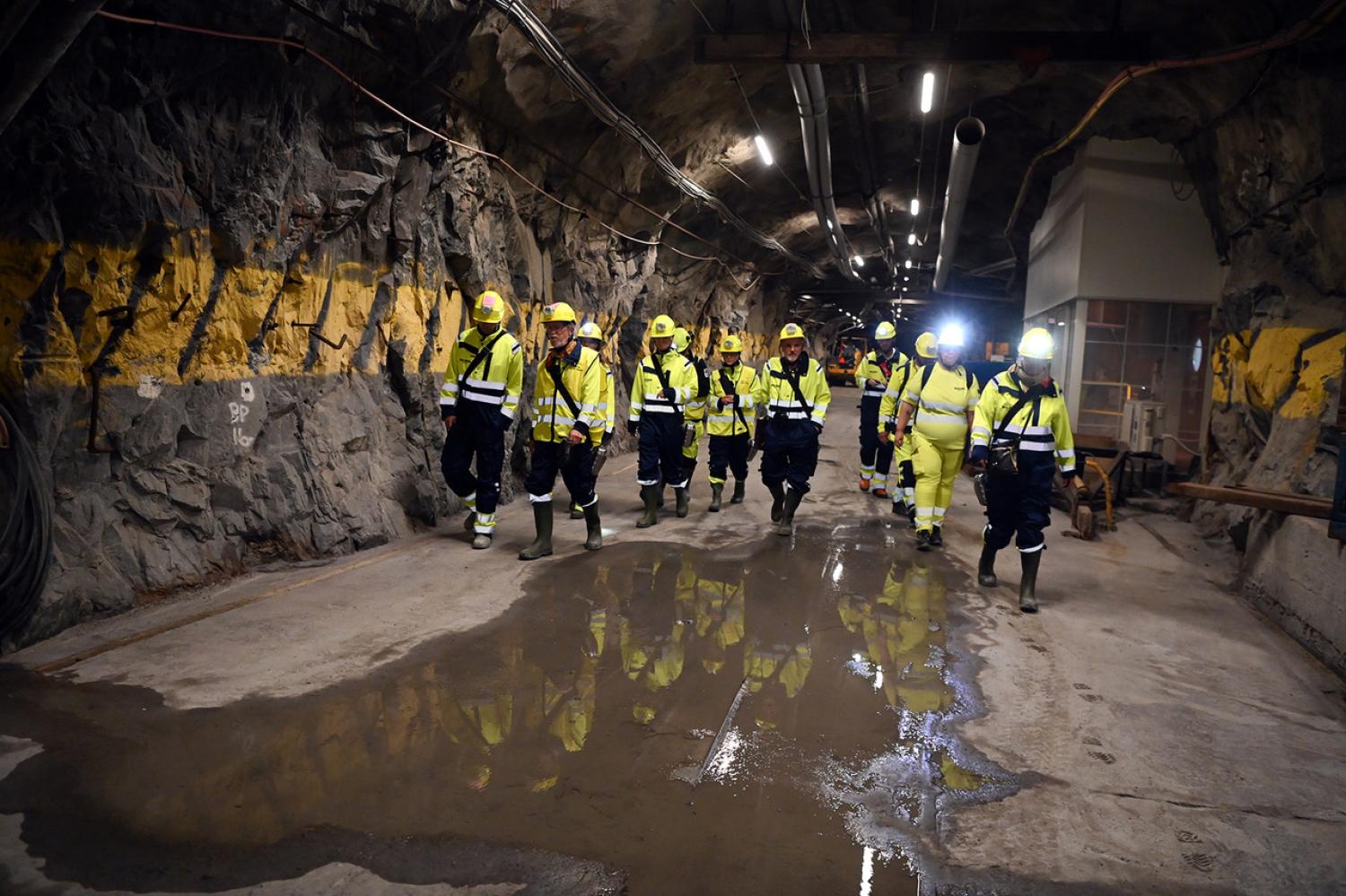 En grupp arbetare i skyddsutrustning går genom en underjordisk tunnel med steniga väggar och våta fläckar på marken.