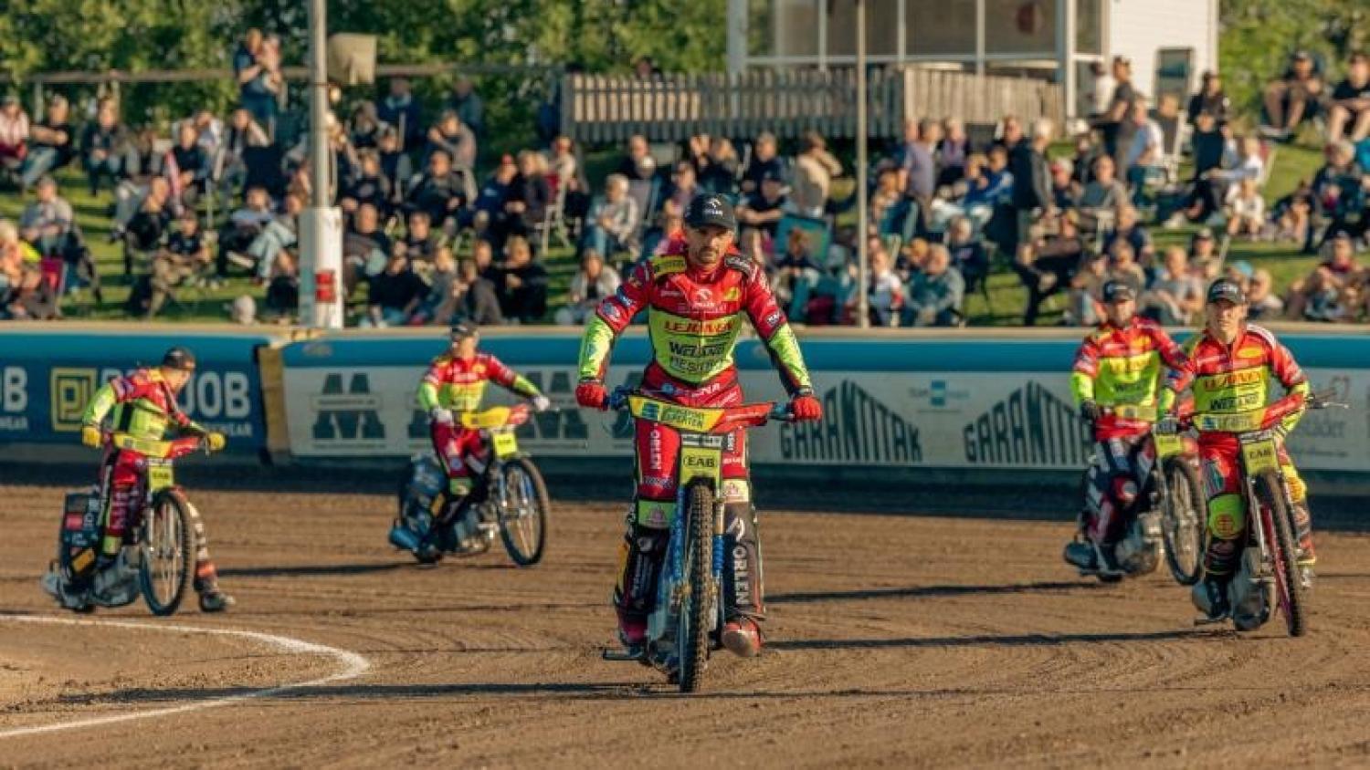 Fem speedwaymotorcyklister i matchande rödgula racingkläder kör på en grusbana, med åskådare som tittar på från läktaren i bakgrunden.