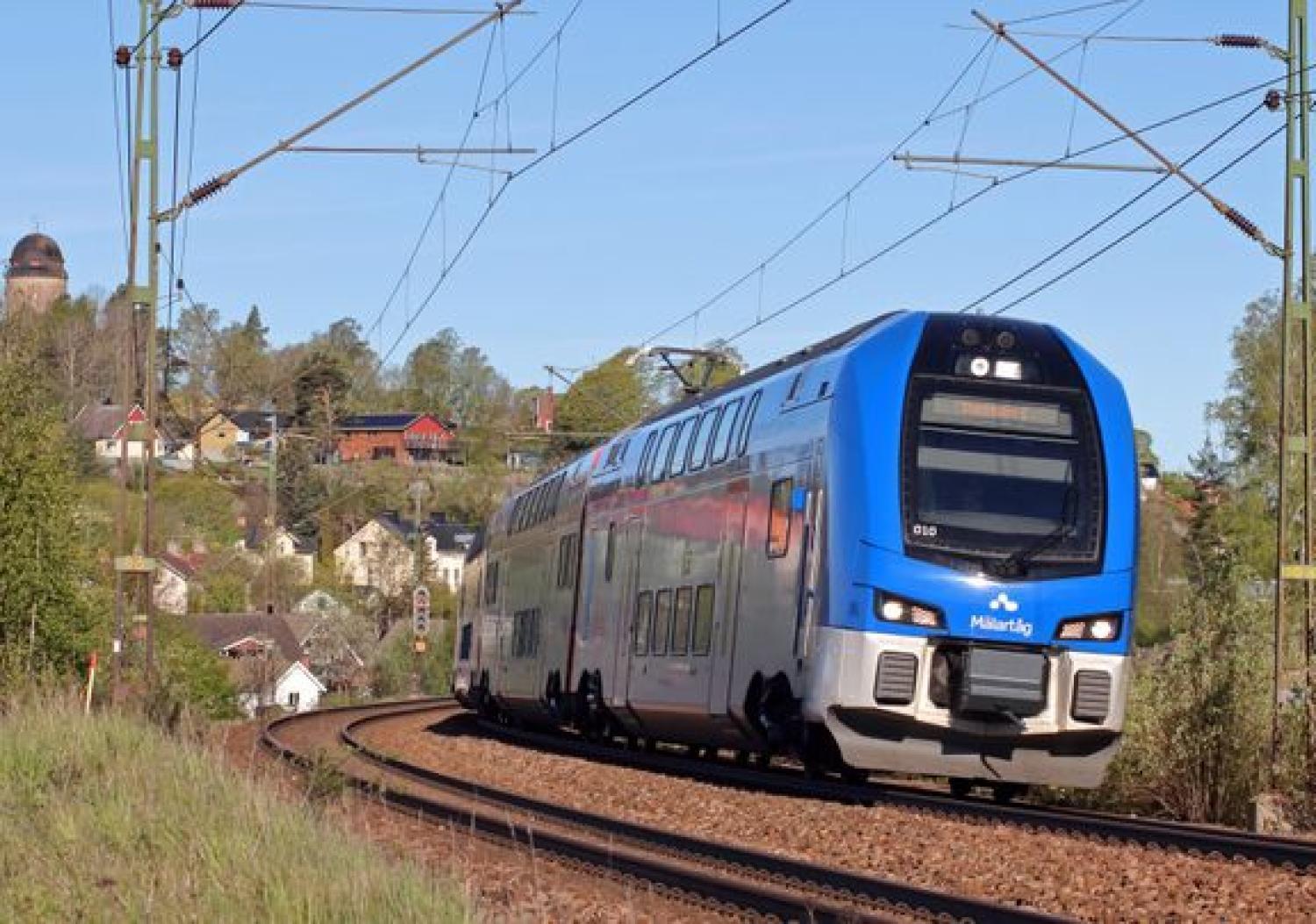 Ett blåvitt dubbeldäckat tåg färdas på en krökt bana genom ett landsbygdsområde med hus och träd i bakgrunden.