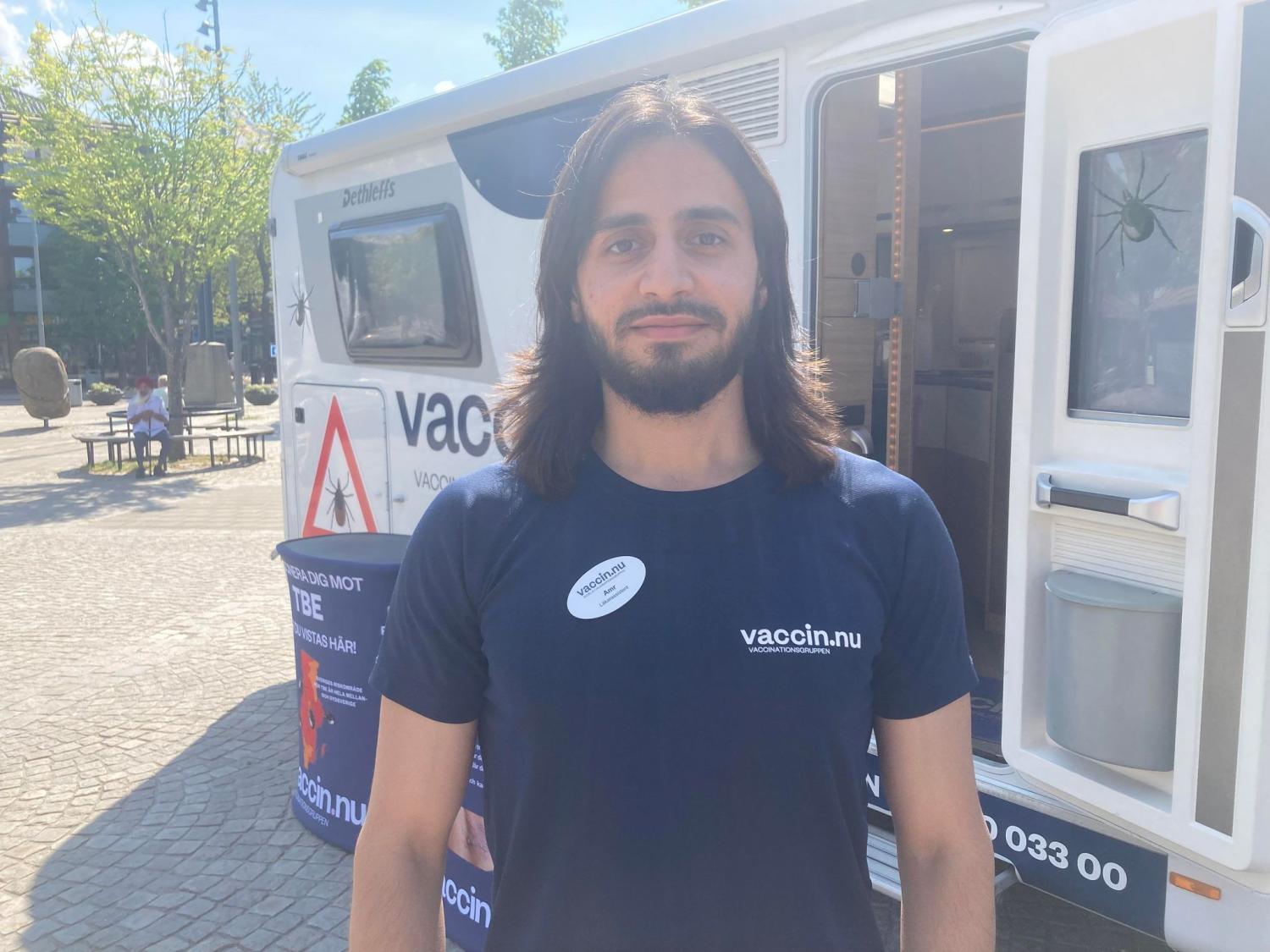 En man med långt hår och skägg står framför en mobil vaccinationsenhet, iklädd marinblå skjorta och namnskylt. Bakgrunden visar ett stenlagt område med några träd och byggnader.