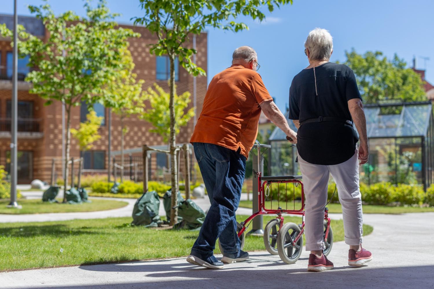 Två äldre personer promenerar utomhus en solig dag; en av dem använder en rullator som stöd. I bakgrunden syns träd och byggnader.