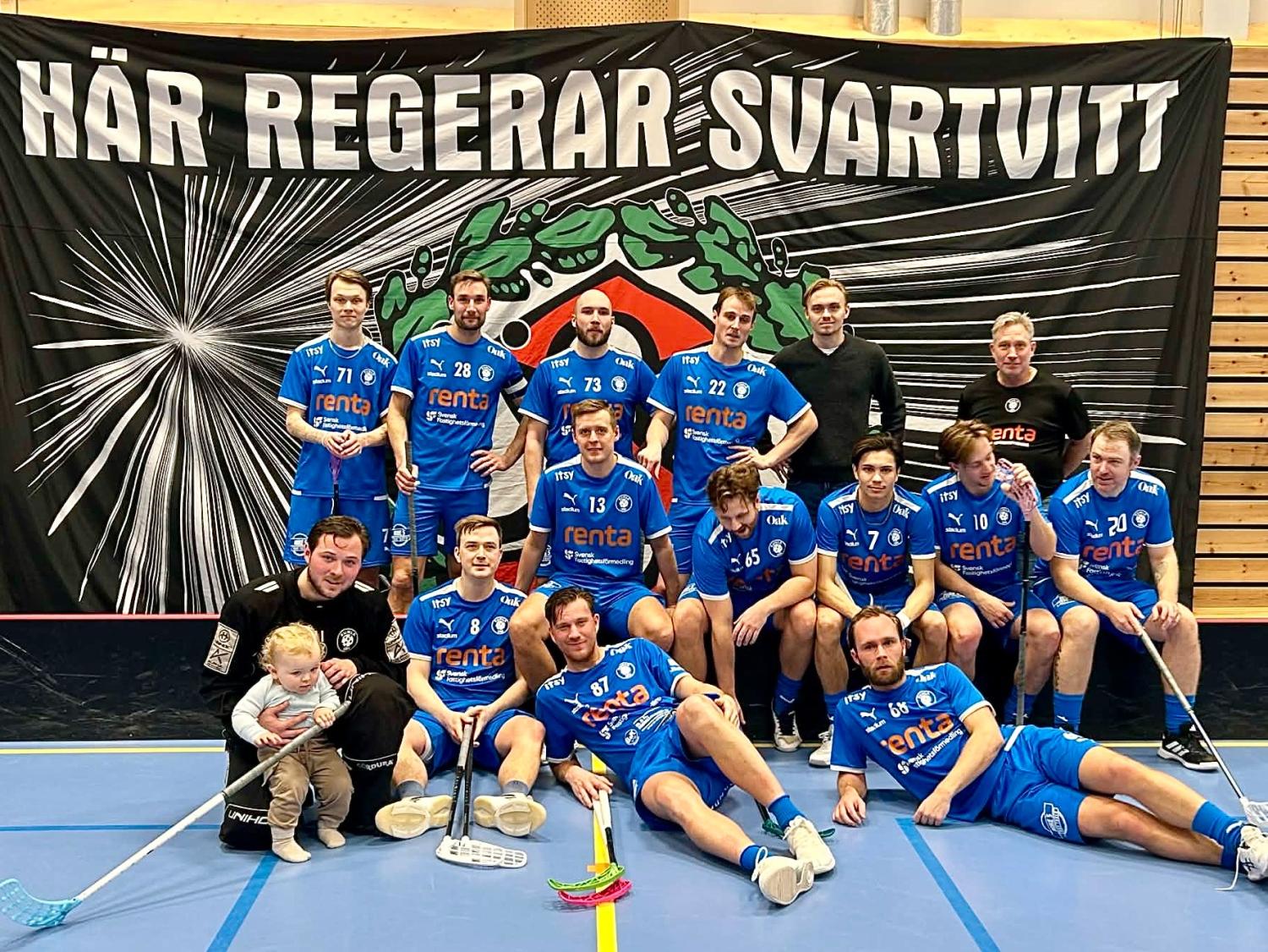 Ett innebandylag för herrar i blåa uniformer poserar inomhus med sina klubbor framför en stor svartvit banderoll. En spelare håller i ett litet barn.