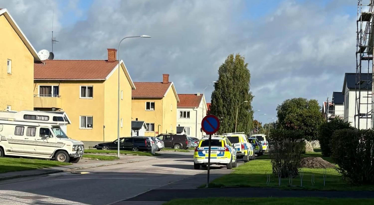 Flera polisbilar står parkerade längs en bostadsgata kantad av gula hus under en delvis molnig himmel.