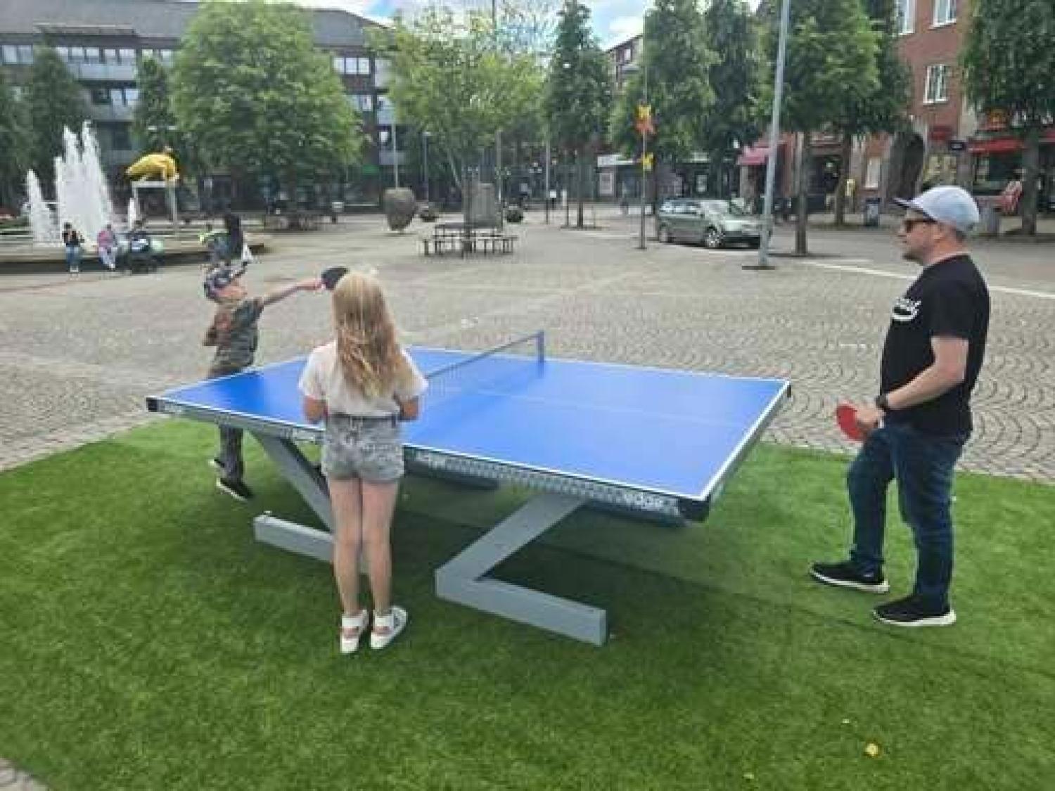 Två barn och en vuxen spelar bordtennis vid ett blått utomhusbord på ett torg med träd, byggnader och en fontän i bakgrunden.