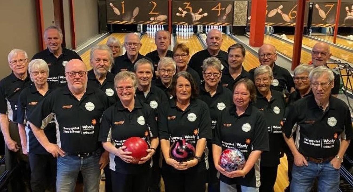 En grupp vuxna i matchande skjortor står tillsammans i en bowlinghall, några håller i bowlingklot och bowlingbanorna syns i bakgrunden.