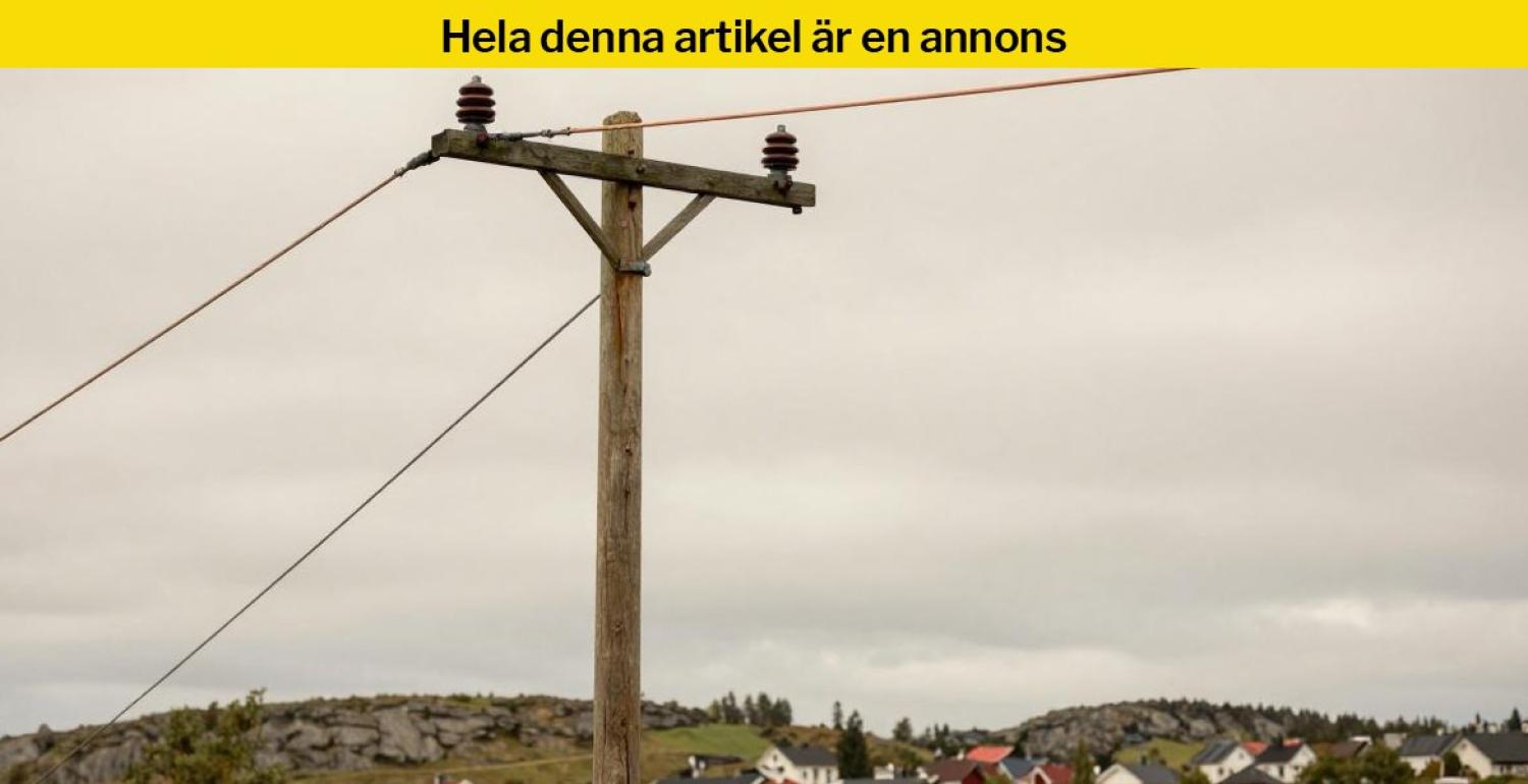 En trästolpe med kraftledningar står framför en molnig himmel, med hus och stenig terräng i bakgrunden.