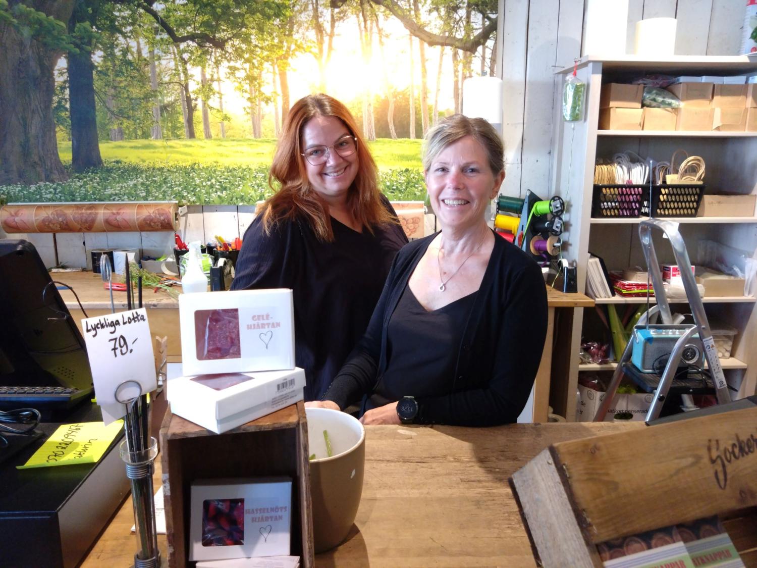 Två kvinnor som står bakom en trädisk i en butik med produkter som visas. En skogsbakgrund syns i bakgrunden.