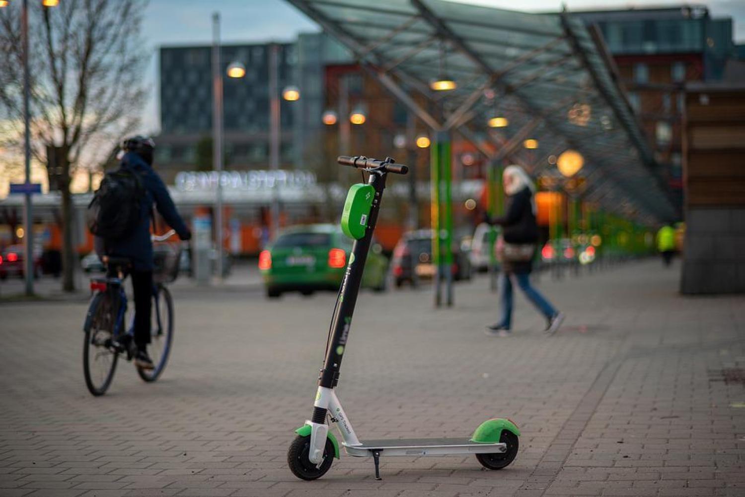 En grön och vit elscooter står parkerad på en trottoar i stan. I bakgrunden ser man människor som går och cyklar under ett glastak.
