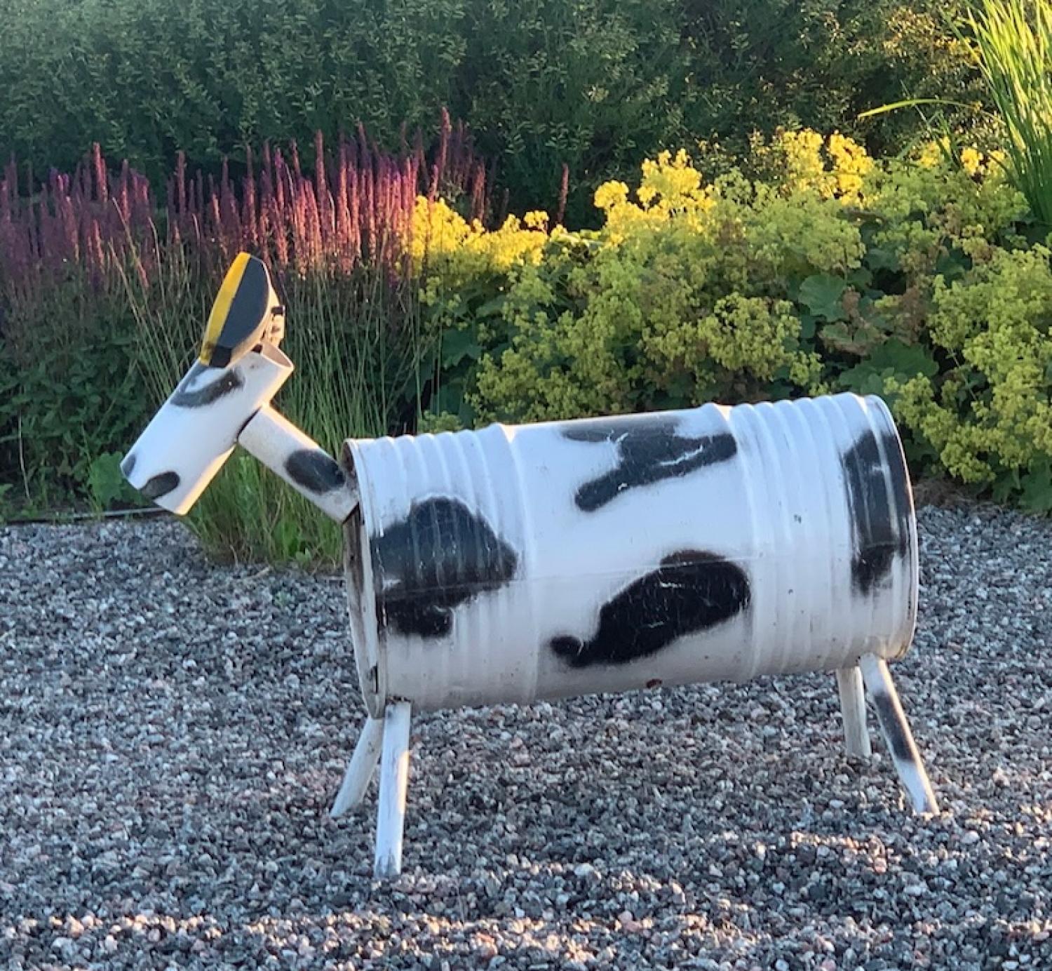 En skulptur av en ko gjord av en målad metalltunna och rör står på grus med gula blommor och grönska i bakgrunden.