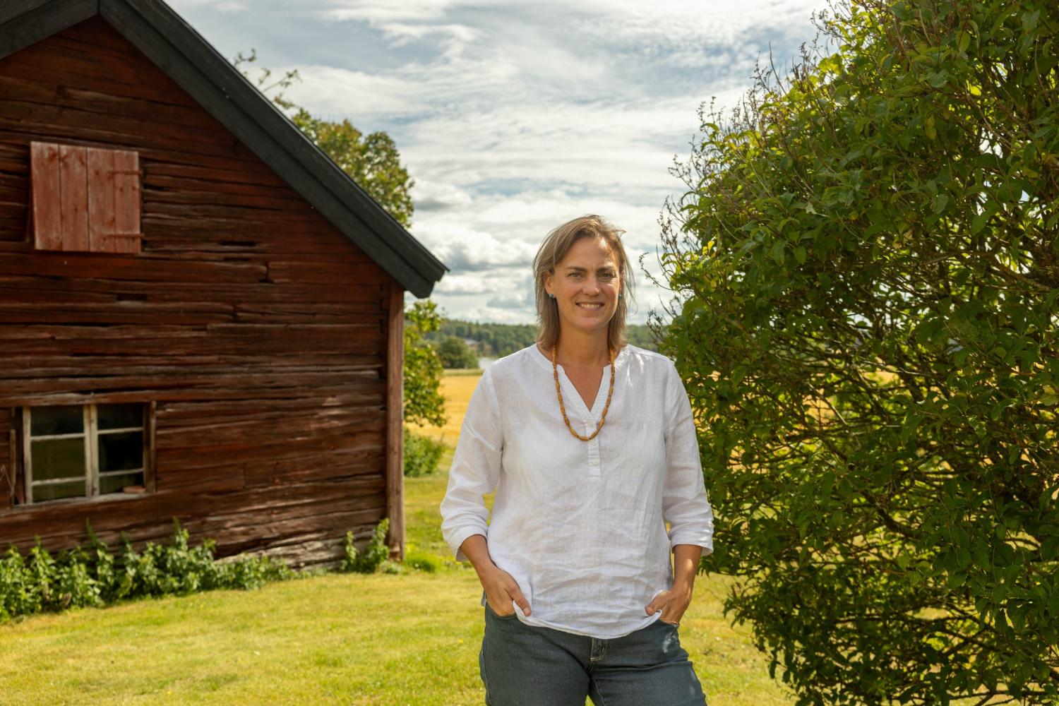 En kvinna i vit skjorta står vid en buske intill en väderbiten träbyggnad en solig dag, med åkrar i bakgrunden.