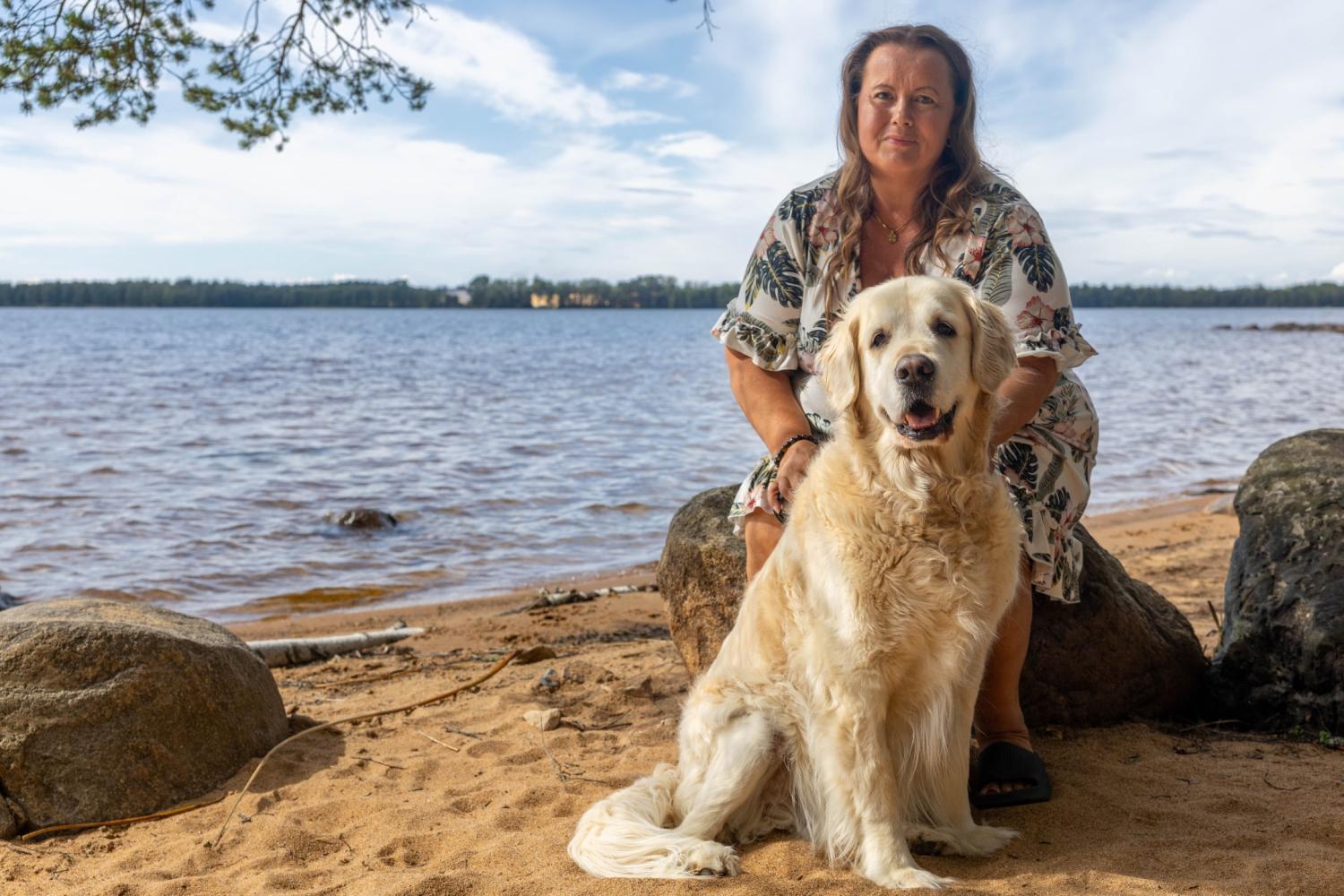 En kvinna i en blommig klänning sitter på en sten vid stranden, med en stor golden retriever som sitter framför henne på sandstranden.